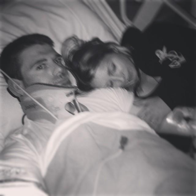 Black and white photo of Dustin Fox in a hospital bed wearing a neck brace while his girlfriend comforts him.